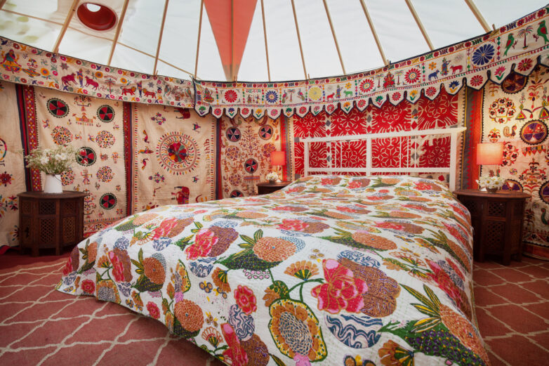 A cosy, colourful yurt bedroom, features patterned quilts, intricate décor, a wooden bed, bedside tables with lamps, fresh flowers in vases, and a striking red geometric rug.