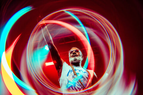 A man wearing sunglasses and a T-shirt raises his fist on stage at Shambala Festival UK, surrounded by swirling, colourful light trails that create a dynamic, energetic effect.