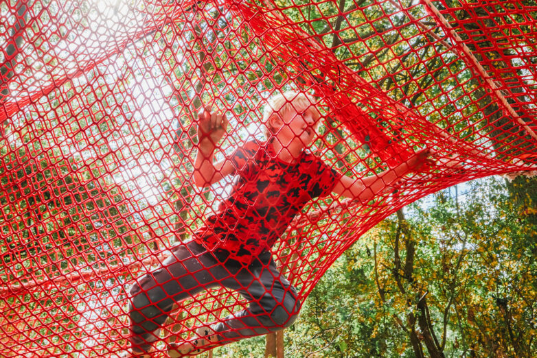 A person with fair hair and a red shirt climbs through a bright red net structure outdoors, surrounded by green trees and sunlight filtering through the leaves.
