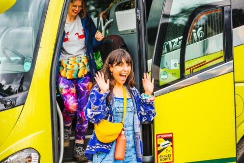 A smiling woman with her hands raised excitedly steps off a yellow bus, followed by another woman. Both wear colourful, casual clothing and bum bags. The bus door is open, showing a safety sticker near the entrance.