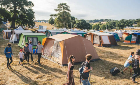 People walk among colourful tents set up on a grassy field at a campsite during the day, with trees and hills in the background under a partly cloudy sky.